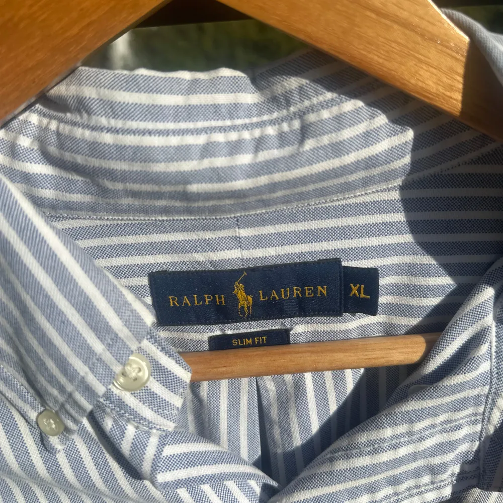 Säljer en snygg och stilren randig skjorta från Ralph Lauren i storlek XL. Skjortan är blå och vit med långa ärmar och en klassisk krage. Den har en broderad logga på bröstet och knappar framtill. Perfekt för både vardag och fest!. Skjortor.