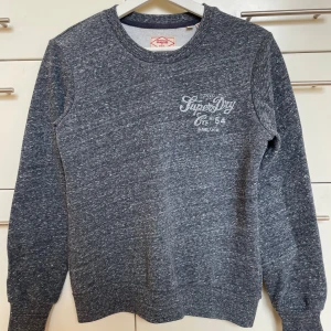 Grå tröja från Superdry - Säljer en supersnygg grå collegetröja från Superdry. Den har en klassisk rund hals och är i ett melerat grått mönster med Superdry:s vita logga på bröstet. Använd en gång - perfekt skick!