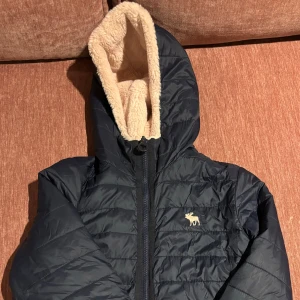 Svart och beige vändbar jacka med huva - Säljer en supermysig vändbar jacka från A&F! Ena sidan är marin och quiltad med en dragkedja och en söt älglogga på bröstet. Den andra sidan är ljusrosa och fluffig. Jackan har en huva och långa ärmar, vilket gör den idealisk för vintern/hösten!