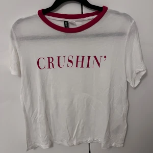 Vit t-shirt med text från Divided - Säljer en vit t-shirt från Divided med röd text 'CRUSHIN'' på framsidan. T-shirten har en röd krage och korta ärmar.