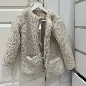 Vit teddyjacka från Ebbe - Mysig vit teddyjacka från Ebbe med dragkedja framtill. Jackan har en fluffig textur och är perfekt för kyligare dagar. Den har långa ärmar och en slät insida för extra komfort.