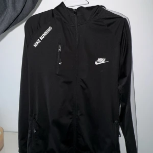 Svart träningsjacka från Nike - Snygg svart träningsjacka från Nike med vit logga och texten 'Nike Running'. Jackan har dragkedja framtill och praktiska fickor. Perfekt för löpning och andra träningsaktiviteter.