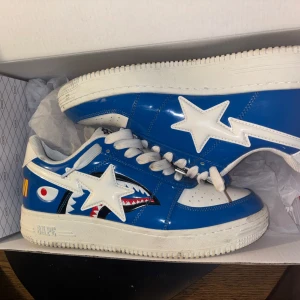 Blå och vita sneakers från Bape - Snygga Bape sneakers i blått och vitt med ikonisk stjärndetalj och hajmönster på sidan. Skorna har snörning och en robust sula, perfekt för en streetwear-look.