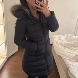 Marinblå dunjacka med pälsdetalj från Moncler - Snygg svart dunjacka från Moncler med pälsdetalj på huvan. Jackan har en figurnära passform och stängs med dragkedja. Perfekt för kalla vinterdagar. Har bara andvänts 1 år köpt i barn storlek 14 år men skulle säga att det är som storlek s priset kan alltid diskuteras samt medföljer mössan med jackan som matchar, kvitton bevis extra skickas med. Köpt för 9500kr