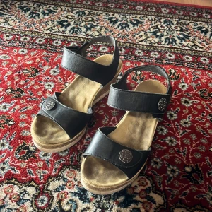 Svarta sandaler från Rieker - Snygga svarta sandaler från Rieker med justerbara remmar och dekorativa detaljer. Perfekta för sommarens äventyr. Bekväma och stilrena med en stabil sula.