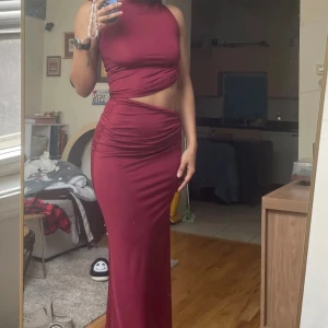 Vinröd tvådelad klänning - Snygg vinröd tvådelad klänning med en ärmlös topp och långkjol. Toppen har en asymmetrisk design med draperade detaljer och kjolen har en smickrande passform. Perfekt för en elegant kvällslook.