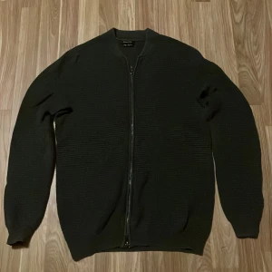Mörkgrön kofta från Massimo Dutti - Snygg mörkgrön kofta från Massimo Dutti med dragkedja framtill. Den har en ribbad struktur och långa ärmar, perfekt för kyligare dagar. Stilren och enkel att matcha med olika outfits.