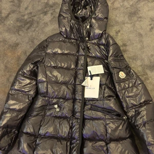 Svart dunjacka från Moncler - Snygg och glansig svart dunjacka från Moncler med huva och dragkedja. Jackan har praktiska fickor med dragkedjor och en klassisk Moncler-logga på ärmen. Perfekt för kyliga dagar.     Storlek, S