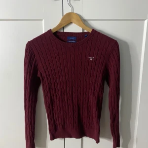 Vinröd kabelstickad tröja från GANT - Säljer en stilren vinröd kabelstickad tröja från GANT i stretchig bomull. Tröjan har en klassisk rund hals och långa ärmar, perfekt för kyligare dagar. En tidlös design som passar till många tillfällen.