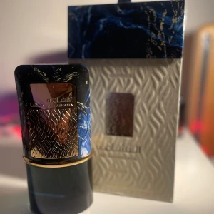 Lattafa Al Nashama edp - Elegant parfym från Al Haramain i en lyxig flaska med svart och blått marmorliknande mönster och gulddetaljer. Förpackningen har en stilren design med en fönsteröppning som visar flaskan. Perfekt för den som söker en sofistikerad doftupplevelse. Köpt nytt och ndast testad.