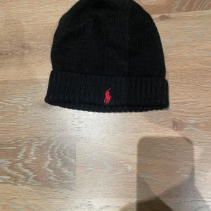 Svart stickad mössa från Ralph Lauren - Snygg svart stickad mössa från Ralph Lauren med röd broderad logga framtill. Perfekt för kyliga dagar med sin ribbade kant som ger en skön passform.