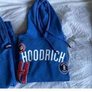 Blå hoodie från Hoodrich och byxor  - Säljer en blå hoodie från Hoodrich med stor vit logga på framsidan och flera detaljer som broderade märken. Hoodien har en klassisk känguruficka och justerbar huva med dragsko. Perfekt för en avslappnad stil.