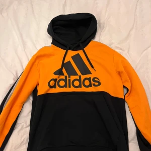 Svart och orange hoodie från Adidas - Säljer en snygg svart och orange hoodie från Adidas med stor logga på framsidan. Tröjan har en klassisk design med huva och långa ärmar. Perfekt för en sportig look eller avslappnade dagar.