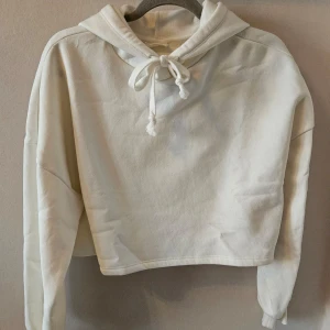 Vit croppad hoodie från Gina Tricot - Säljer en vit croppad hoodie från Gina Tricot. Väldigt mjukt mysigt tyg på insidan. Den har en justerbar huva med knytband och långa ärmar. Perfekt för en avslappnad stil. Aldrig använd men lapp ej kvar.
