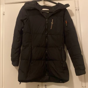 Svart dunjacka från Sail Racing - Snygg svart dunjacka från Sail Racing med dragkedja och huva. Perfekt för kyliga dagar med sin långa design och vadderade stil. Jackan har en logotyp på bröstet och en praktisk ficka på ärmen.