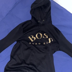 Svart hoodie från Hugo Boss - Snygg svart hoodie från Hugo Boss med guldtryck på framsidan. Perfekt för en stilren och bekväm look. Den har en klassisk Luva med dragskedja och en stor ficka framtill.