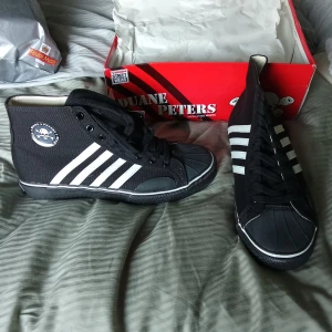 Svarta och vita DP Canvas Hi Top sneakers - Snygga svarta och vita DP Canvas Hi Top sneakers med klassiska ränder på sidan. Skorna har en hög modell med snörning och en gummisula i ljusbrun färg. Perfekta för en cool och avslappnad stil.