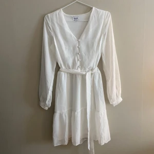Vit klänning med knappar och bälte - Elegant vit klänning med v-ringning och knappar framtill. Perfekt för vår och sommar och student. Klänningen har bara används en gång så i nyskick!