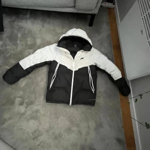 Svart och vit dunjacka från Nike - Snygg och varm dunjacka från Nike i svart och vitt. Jackan har en huva och dragkedja framtill. Perfekt för kyliga dagar.