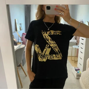 Svart t-shirt med guldtryck - Snygg svart t-shirt med guldtryck framtill. Kortärmad och i normal passform. Tröjan är i ett mycket fint skick! 