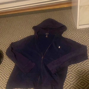 Mörkblå hoodie från Polo Ralph Lauren - Säljer en snygg mörkblå hoodie från Polo Ralph Lauren med dragkedja och huva. Den har en liten broderad logga på bröstet och är perfekt för kyliga dagar. Passar både till vardags och avslappnade tillfällen. Kan gå ner i pris vid snabb affär.