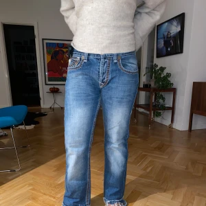 Lågmidjade true religion jeans - Snygga lågmidjade blå jeans från True religion med broderade detaljer på bakfickorna. De har en klassisk femficksdesign och en bekväm passform. Perfekta för en avslappnad stil. Något slitna nertill och är lagade i grenen annars i fint skick.