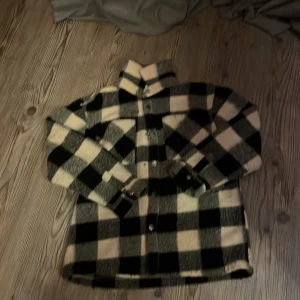 Rutig jacka från Lindex - Säljer en snygg rutig jacka från Lindex i svart och beige. Jackan har långa ärmar och knappar framtill. Perfekt för höst och vår med sitt mjuka material av återvunnen polyester.