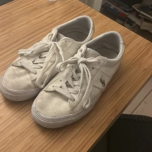Vita sneakers från Ralph Lauren - Säljer ett par vita sneakers från Ralph Lauren med snörning och klassisk design. Skorna har en broderad logotyp på sidan och är perfekta för en avslappnad stil.