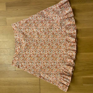 Blommig kjol från NA-KD - Söt blommig kjol från NA-KD i härliga nyanser av rosa, orange och grön. Kjolen har en volangdetalj längs nederkanten som ger en lekfull touch. Perfekt för somriga dagar!