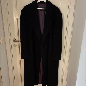 Lång svart överrock - Lång stilig överrock i ull. Smått luggsliten från den förra ägaren men inget markant. Perfekt nu i kallare väder. (Längd 114cm, axel 53cm, ärm 58cm, bröst 53cm)