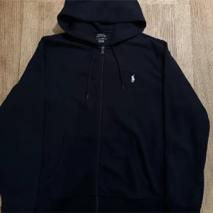 Svart hoodie från Ralph Lauren - Snygg svart hoodie från Ralph Lauren med dragkedja och klassisk logga på bröstet. Perfekt för en avslappnad stil. Den har en justerbar huva och praktiska fickor framtill.