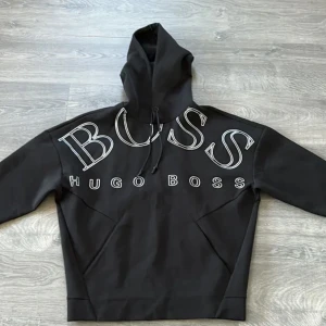 Svart hoodie från Hugo Boss - Snygg svart hoodie från Hugo Boss med stor logga på bröstet. Perfekt för en stilren och avslappnad look. Den har en klassisk huva med dragsko och en bekväm passform. Helt nytt 
