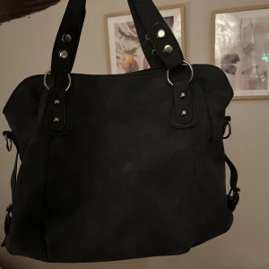 Svart axelväska med nitar - Snygg svart axelväska med dekorativa nitar på handtagen. Väskan har en stilren design och är perfekt för att bära med sig det nödvändigaste. Den har robusta handtag och en praktisk storlek för dagligt bruk. Köpt här på Plick men aldrig kommit till användning av mig