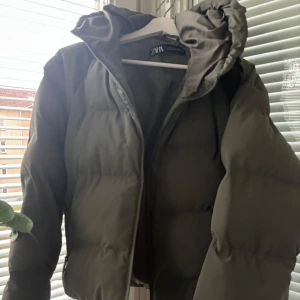 Grön pufferjacka från Zara - Säljer en snygg grön pufferjacka från Zara. Den är perfekt för kalla dagar med sin tjocka vaddering och har en praktisk huva. Jackan har en dragkedja framtill och är i mycket bra skick. Perfekt för både höst och vinter! Storlek S men skulle säga att den också funkar för M.