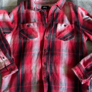 Stussy - Säljer en snygg röd och svart rutig skjorta från Stussy i storlek M. Skjortan har långa ärmar och knappar framtill samt på bröstfickorna. Perfekt för höst och vinter med sitt mjuka och bekväma material. Passar både till vardags och festliga tillfällen!