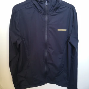Mörkblå vindjacka med huva från A-Z - Säljer en stilren mörkblå vindjacka från A-Z. Den har en praktisk huva och dragkedja framtill. Jackan är använd ett fåtal gånger och är i mycket bra skick!