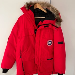 Canada goose jacka röd - En röd Canada Goose-jacka i högkvalitativt material som håller dig varm under kalla vinterdagar. Jackan har ett trasigt blixtlås men är annars i gott skick och ett utmärkt fynd för den som är beredd att laga det. Perfekt för både funktion och stil.