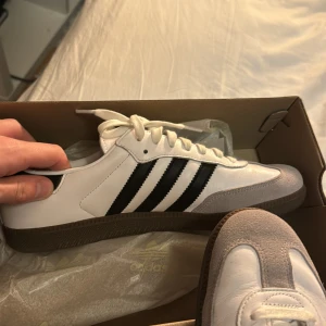 Adidas Samba i vitt och svart - Säljer ett par klassiska Adidas Samba sneakers i vitt och svart. De har den ikoniska tre-randiga designen och en snygg brun sula. Perfekta för både vardag och fest! Skorna är i mycket bra skick och kommer i originalkartong.