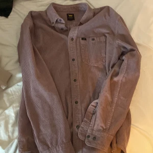 Lila manchester skjorta från Lee - Säljer en snygg lila manchester skjorta från Lee i avslappnad passform. Skjortan har långa ärmar och knappar framtill samt en bröstficka. Perfekt för höst och vinter, både stilren och bekväm. Passar lika bra till vardags som till festliga tillfällen!