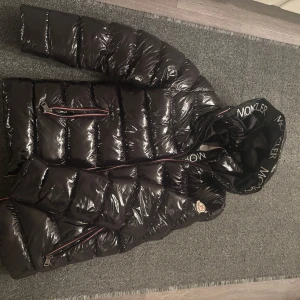 Svart pufferjacka från Moncler - Säljer en snygg svart pufferjacka från Moncler i mycket bra skick. Jackan har en glansig finish och är perfekt för vintern. Den har en dragkedja framtill och Moncler-logga på ärmen. Huvan är fodrad och har Moncler-tryck. Perfekt för att hålla sig varm och stilren i kylan!