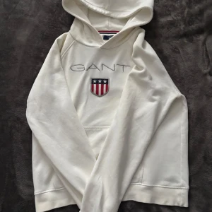 S Vit hoodie från GANT  - Säljer en snygg vit hoodie från GANT med deras klassiska logga och ett broderat emblem på bröstet. Den är i mycket bra skick och perfekt för både höst och vår. Hoodien har en skön passform och är gjord i mjukt material. Perfekt för en casual look eller mysiga dagar hemma!