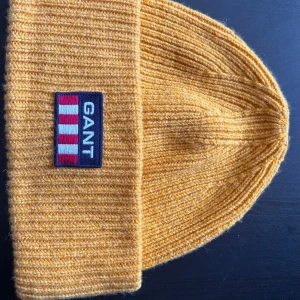 Gul stickad mössa från GANT - Säljer en snygg gul stickad mössa från GANT. Den har en snygg design och ett broderat märke framtill. Perfekt för att hålla sig varm under höst och vinter. Mössan är i mycket bra skick och passar de flesta.