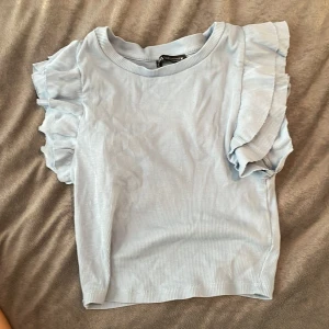 Ljusblå topp med volangärmar från Zara - Säljer en superfin ljusblå topp från Zara i storlek S. Den ehar volangdetaljer på ärmarna som ger en söt touch! Toppen är i ribbat material och känns bekväm. Den riktiga färgen är på bild 3 och 4