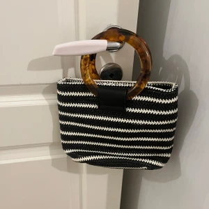 Randig handväska med runda handtag - Säljer en superfin randig handväska i svart och vit. Den har unika runda handtag i brun plast som ger en cool retro-look. Perfekt för att piffa upp vilken outfit som helst! 🖤🤍