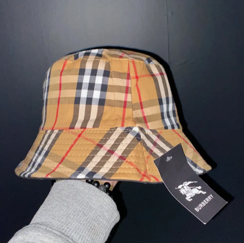 Säljer en snygg bucket hat från Burberry i deras klassiska rutiga mönster. Hatten är i beige, svart och röd med en bred kant. Perfekt för att ge din outfit en stilren touch. Helt ny med tag kvar!. Accessoarer.