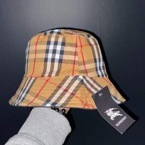 Säljer en snygg bucket hat från Burberry i deras klassiska rutiga mönster. Hatten är i beige, svart och röd med en bred kant. Perfekt för att ge din outfit en stilren touch. Helt ny med tag kvar!