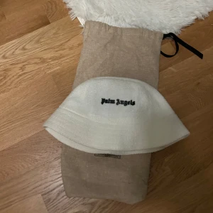 Vit bucket hat från Palm Angels - Säljer en stilren vit bucket hat från Palm Angels. Hatten har en broderad logotyp i svart framtill och är tillverkad i ett mjukt material. Perfekt för att ge en cool touch till din outfit.