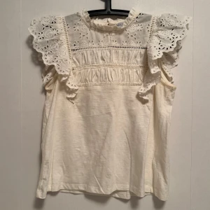 Vit blus från Holly & Whyte - Charmig vit blus från Holly & Whyte med vackra spetsdetaljer och volanger på axlarna. Blusen har en rund hals och knäppning bak. 