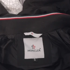 Svart jacka från Moncler - Säljer en stilren svart jacka från Moncler med dragkedja och knappar. Jackan har en klassisk design med Moncler-logga på bröstet och en huva för extra skydd. Perfekt för kyligare dagar.