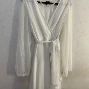 Vit omlottklänning med prickar - Säljer en elegant vit omlottklänning med prickigt mönster. Klänningen har långa ärmar och ett knytband i midjan för en smickrande passform. Perfekt för en stilren look.